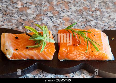 Eingelegte Lachsstücke auf einem Holzbrett auf Marmorboden, verziert mit Salbei- und Rosmarinblättern Stockfoto