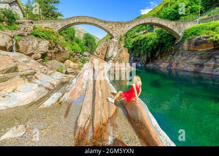 Frau, die an der alten römischen Steinbrücke sitzt: Ponte dei Salti über dem Verzasca-Fluss. Verzasca Tal bei der Stadt Lavertezzo. Berühmtes Wahrzeichen für Stockfoto