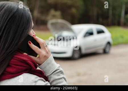 Nahaufnahme einer Latina-Frau, die auf einem Handy mit ihrem kaputten Auto im Hintergrund spricht Stockfoto