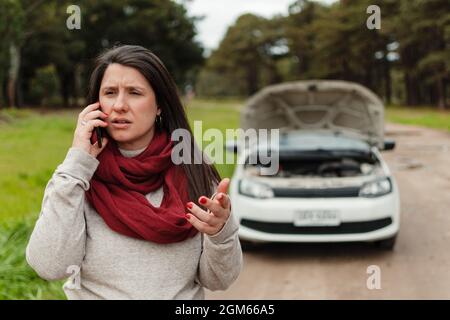 Nahaufnahme einer Latina-Frau, die am Handy spricht und gestikuliert. Ihr kaputtes Auto im Hintergrund Stockfoto