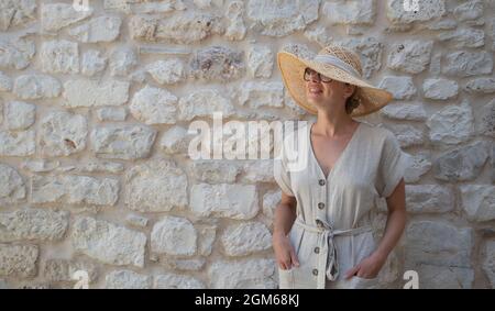 Portrait einer schönen fröhlichen blonden Frau, die ein einteiliges Sommerkleid und Sommerhut trägt und vor einer alten mittelalterlichen Steinmauer steht. Sommer Stockfoto