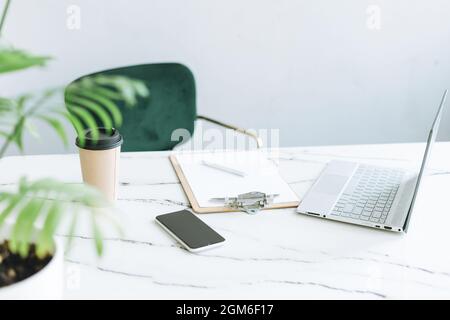 Arbeitsplatz mit Papiertasse, Notizen, offenem Laptop und Smartphone auf weißem Marmortisch im hellen, modernen Bürozimmer, Geschäftskonzept Stockfoto