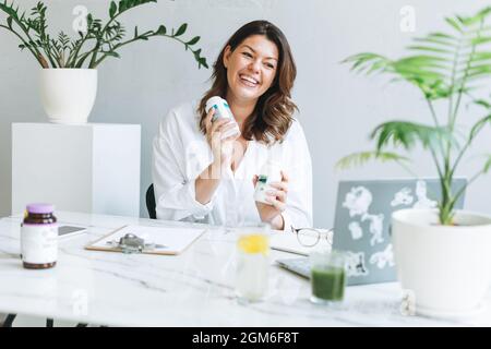 Junge lächelnde Brünette Frau Ernährungsberaterin plus Größe in weißem Hemd arbeiten am Laptop auf dem Tisch mit Haus Pflanze in hellen modernen Büro. Mitteilung des Arztes Stockfoto