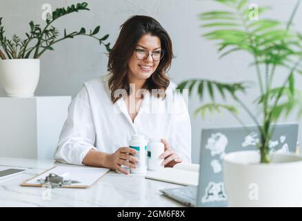 Junge lächelnde Brünette Frau Ernährungsberaterin plus Größe in weißem Hemd arbeiten am Laptop auf dem Tisch mit Haus Pflanze in hellen modernen Büro. Mitteilung des Arztes Stockfoto