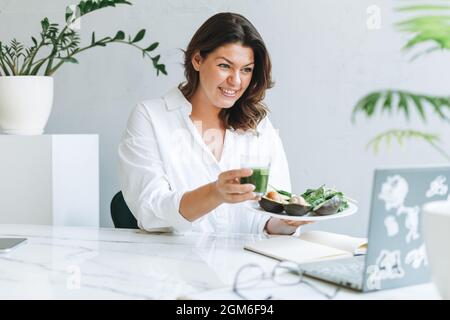 Junge lächelnde Brünette Frau Ernährungsberater Arzt plus Größe in weißem Hemd mit grünen gesunden Lebensmitteln im modernen hellen Bürozimmer. Arzt kommuniziert Stockfoto