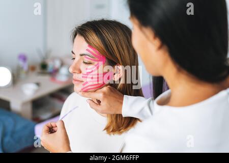 Junge schöne Brünette Frau mit langen Haaren Arzt Kosmetologe macht Gesicht Taping für Patienten im Büro. Junge Frauen führen Anti-Aging-Verfahren mit durch Stockfoto