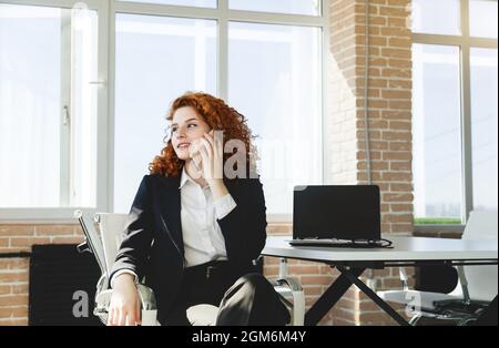 Porträt einer schönen erfolgreichen jungen Geschäftsfrau mit roten Lockenhaaren in einem Büroinnenraum, die an einem Tisch sitzt und am Telefon spricht Stockfoto