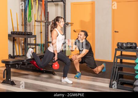 Frau macht Ausfallschritte mit einer Kugelhantel mit Hilfe eines Trainers im Fitnessstudio. Stockfoto