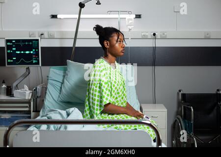 Junge afroamerikanische Person, die mit Fieber, Krankheit und Krankheit auf dem Krankenhausbett sitzt. Kranker Teenager mit Oximeter zur Hand, medizinischen Geräten und Herzfrequenzsensor zur Genesung Stockfoto