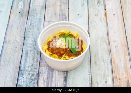 Schüssel mit Nudeln mit Bolognese-Sauce, Käse und Basilikum in Hauslieferbehälter auf Holzhintergrund Stockfoto