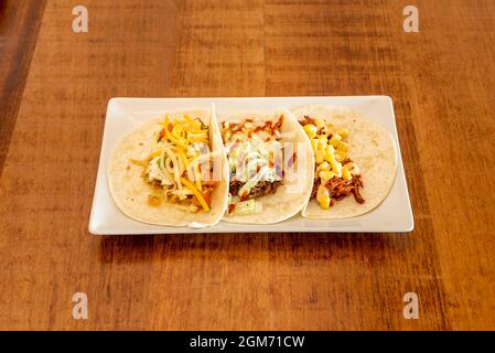 Weizenmehl-Tortilla-Tacos mit verschiedenen Füllungen, geriebenem Käse, zerrissenen Fleisch, süßem Mais auf Holztisch Stockfoto