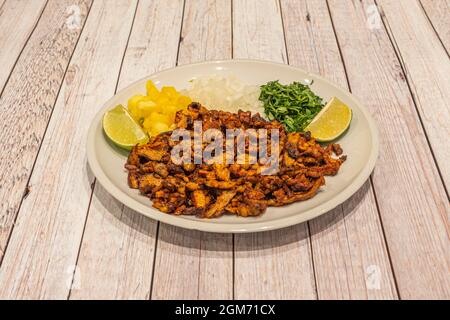 Teller mit Zutaten von Tacos al Pastor. Gehackte Ananas, Gisada-Fleisch, gehackte Zwiebel und Koriander mit zwei Limettenstücken auf einem weißen Teller Stockfoto