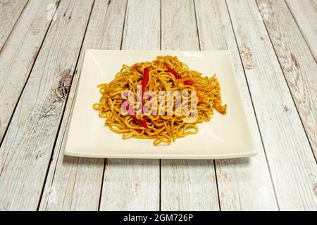 Sautierte Nudeln mit Gemüse und Streifen roter Paprika mit Sojasauce auf weißem quadratischem Teller Stockfoto