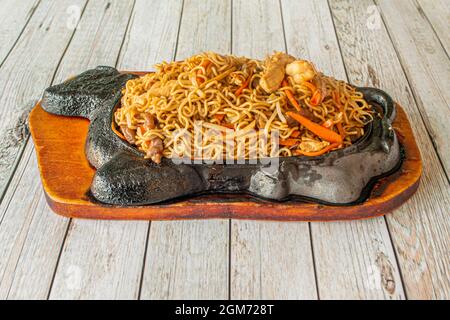 Chinesische Spezialitäten-Nudeln, die mit Garnelen, Rind- und Hühnerfleisch, Gemüse und Karotten auf einem feurigen gusseisernen Tablett anbraten Stockfoto