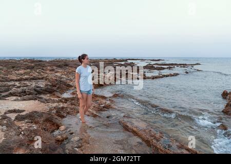 Teenager-Mädchen, das bei Sonnenuntergang auf einer Klippe am Meer steht, trägt ein hellblaues T-Shirt, Jeans-Shorts und schaut weg. T-Shirt-Modell Stockfoto