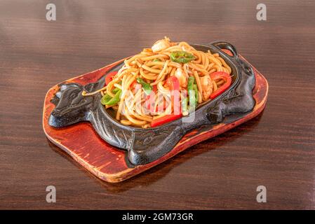 Gebratene udon-Nudeln mit Gemüse und geschälten Garnelen, die von einem chinesischen Koch gekocht und auf einem kochenden gusseisernen Tablett serviert werden Stockfoto