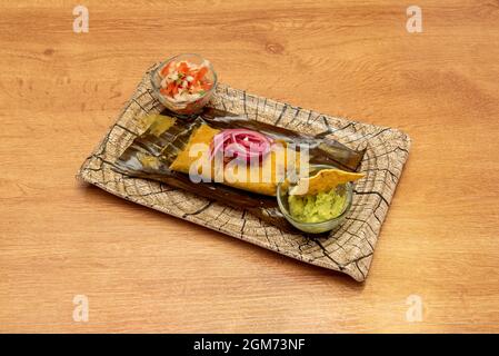 Super mexikanisches Tamale gefüllt mit Cochinita Pibil Fleisch mit Guacamole mit Tortilla Chips und Pico de gallo auf einem Teller ähnlich einem Holzbalken Stockfoto