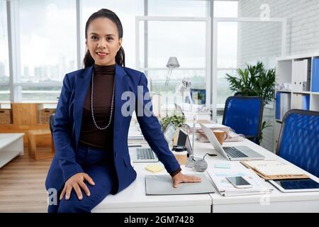 Lächelnde, elegante Geschäftsfrau in blauem Anzug, die sich auf ihren Schreibtisch lehnt und die Kamera anschaut Stockfoto