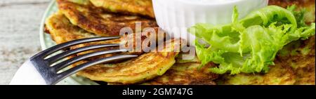 Banner mit Krapfen aus Zucchini mit saurer Sahnesauce und grünem Gemüse. Gemüsepfannkuchen aus Zucchini mit grünem Salat auf weißem Holzhintergrund. Stockfoto