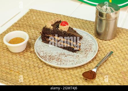 Portion Schokoladenkuchen mit drei Lagen Biskuitkuchen und gefüllt mit Kaffeesahne mit Milch, braunem Zucker und Kakaopulver auf Rattan-Tischtuch Stockfoto