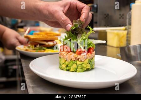 Die Hände des Küchenchefs beendeten die Montage einer Lachstartare mit Avocado und gehackten Kirschtomaten auf dem metallenen Küchentisch auf einem weißen Tablett Stockfoto