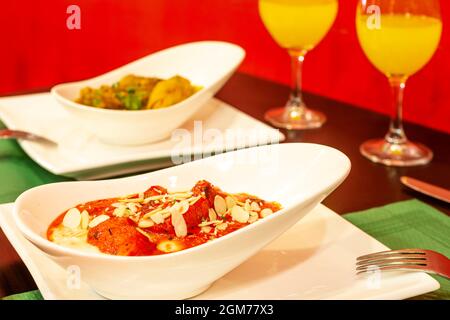 Weiße längliche Schüssel mit Chicken Tikka Masala-Fruchtfleisch mit Mandelscheiben, serviert in einem Restaurant mit Gläsern Orangensaft. Stockfoto