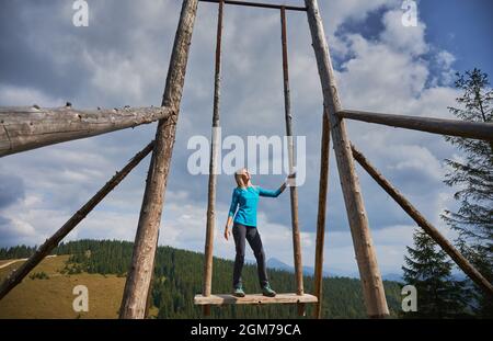 Vor dem Hintergrund des bewölkten Himmels und der Berge entspannen sich die Wanderinnen, die auf einem Sitz mit großer Holzschaukel stehen und sich mit geschlossenen Augen in der warmen Sonne sonnen. Konzept von Reisen, Freiheit und Natur. Stockfoto