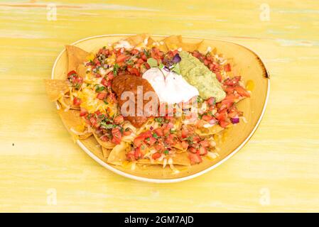 Gelbes Tablett überfüllt mit mexikanischen Mais-Nachos mit Guacamole, Frischkäse, Chili con Carne und geschmolzenem Cheddar-Käse auf gelbem Tisch Stockfoto