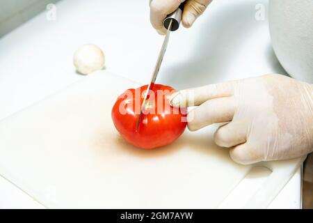 Die Hände des Küchenchefs glänzen mit Latex und schneiden eine reife Tomate auf ein weißes Teflonbrett Stockfoto