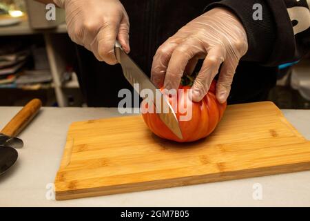 Küchenhände mit Plastikhandschuhen, die eine große reife Tomate auf einem Bambusbrett hacken Stockfoto