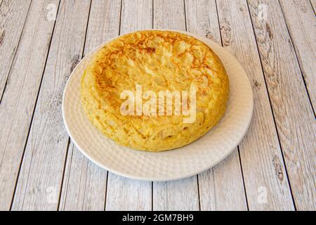 Spanisches Omelett mit Kartoffeln und Eiern mit einer beträchtlichen Dicke auf einem weißen Teller und einem hellen Holztisch Stockfoto