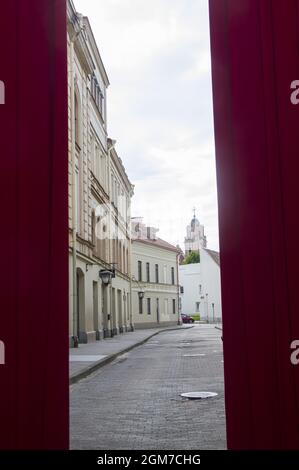 Konzeptionelles Foto der Straße mit Kirche hinter rotem Vorhang in der Altstadt. Architektur von weißen Vintage-Häusern, Apartments mit Fenstern, Balkonen, Boutiquen gegen den bewölkten Himmel im Zentrum von Vilnius Stockfoto