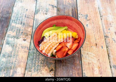 Tropischer Salat mit Erdbeeren, gehackter Mango, bolivianischen Avocados und gekochten galicischen Flusskrebsschwänzen, serviert in einer hübschen roten Schüssel auf einem Holztisch Stockfoto