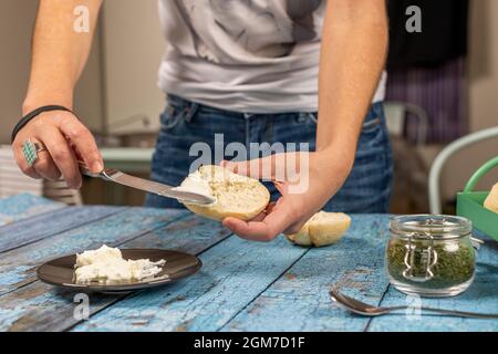 Hände einer jungen Frau in Jeans, die frischen Käse auf frisch gebackenen Muffins verteilt Stockfoto