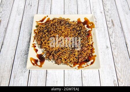 Sautierte Nudeln mit Schweinefleisch, Yakisoba-Sauce und einer großen Menge Mohnsamen und Sesamsamen auf einem quadratischen Teller. Stockfoto