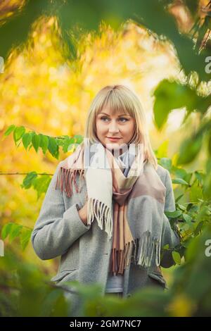 Portrait der blonden Frau mit einem kurzen Haarschnitt geht in einem grauen Wollmantel und einem karierten Schal durch den Wald. Wandern in der Natur im Herbstwald Stockfoto