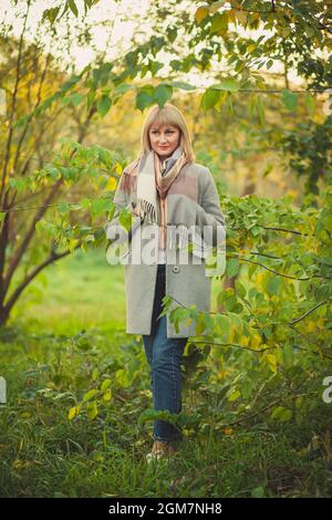 Eine blonde Frau mit einem kurzen Haarschnitt geht in einem grauen Wollmantel und einem karierten Schal durch den Wald. Wandern in der Natur im Herbstwald Stockfoto