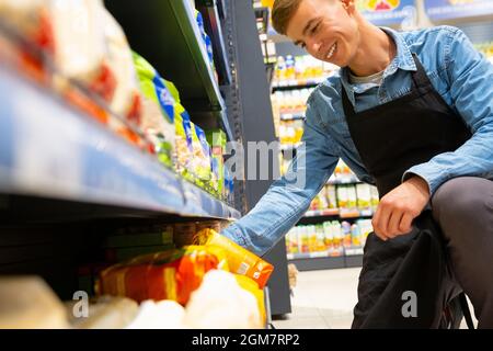 Ein Mitarbeiter mit lächelndem Gesichtsausdruck sitzt auf einem Knie im Geschäft und trägt eine Schürze und überprüft das Verfallsdatum der Cerealien. Stockfoto