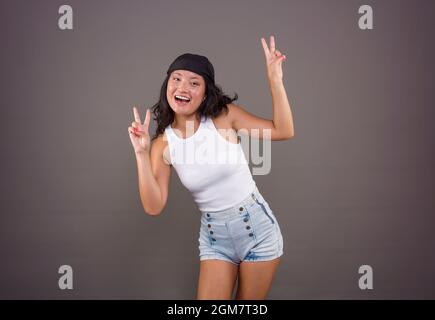 Hübsches chinesisches Mädchen mit Bandana und Shorts, das das Friedenszeichen oder Siegeszeichen macht, in lustiger Pose und Ausdruck Stockfoto