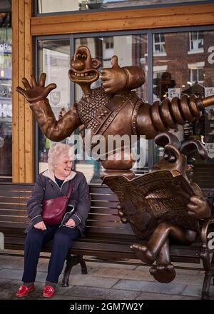 Die Wallace und Gromit Statue vor der Markthalle im Stadtzentrum in Preston, Lancashire, Großbritannien. Ein Passant sitzt auf der Bank neben Gromit Stockfoto