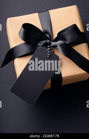 Overhead-Ansicht auf Geschenkbox mit schwarzer Schleife und Anhänger auf schwarzem Hintergrund. Stockfoto
