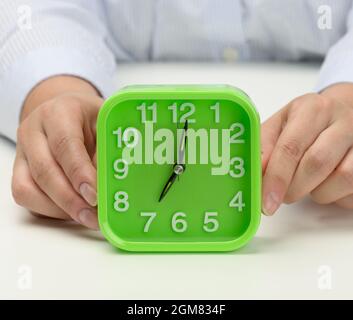 Die Hand der Frau hält einen grünen quadratischen Wecker, die Uhr zeigt sieben Uhr morgens an. Wachen Sie früh auf, und beginnen Sie den Tag Stockfoto