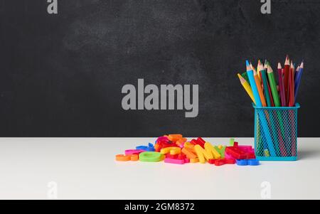 Metallglas mit Stiften, Bleistiften und Filzstiften und Scheren auf dem Hintergrund einer leeren schwarzen Kreidetafel, weißem Tisch, Kopierraum Stockfoto