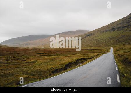 Färöer-Inseln, Streymoy. Spektakuläre Aussicht auf gelbe und braune Hügel mit leerer Straße. Moody nebliger Himmel Stockfoto