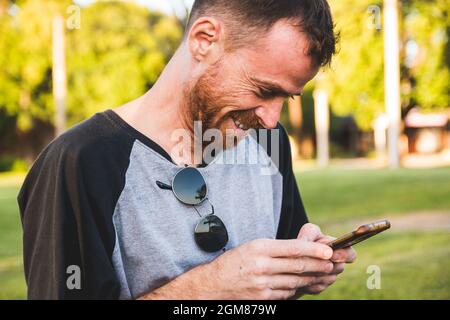Junger attraktiver kaukasischer Mann lächelt und schreibt am Telefon in einem Park Stockfoto