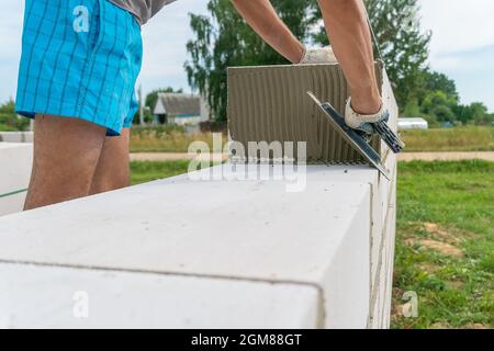 Der Baumeister verwendet eine gekerbte Kelle, um das Zementgemisch aus nächster Nähe auf die belüftelten Betonblöcke aufzutragen. Konzept des Aufbaus eines Privathauses Stockfoto