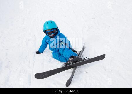 Kleinkind Junge in Helm, Brille, Ski und blauen Overalls im Skigebiet. Skikurs für Kinder an einer Alpinschule. Fiel auf den Schnee. Wintersport Stockfoto