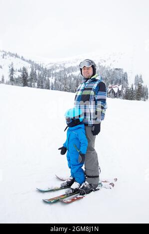 Kleinkind Junge in warmen blauen Overalls, Helm lernt Skifahren mit seinem Vater während der Winterferien in verschneiten Bergen. Sportausbildung, Pflege. Kalter Tag. usef Stockfoto