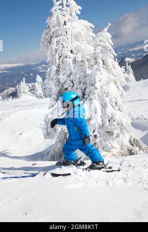 Kleinkind Junge in Helm, Brille und blauen Overalls Skifahren in schönen schneebedeckten Bergen. Aktive Winterunterhaltung für Kinder. Skikurs an einem al Stockfoto