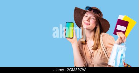 Sehr froh, dass die Touristenfrau mit Hut und Sonnenbrille auf der Kamera posiert und Handy, medizinische Maske und Reisepass in der Hand hält. Stockfoto